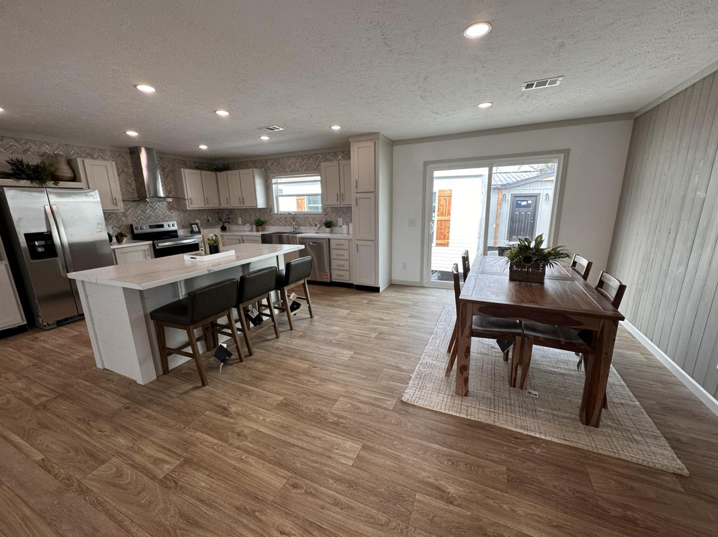 Modern modular home showcase featuring the APH 1662C 819 SQ FT show home and expansive double-wide models, with white vertical board and batten siding, dark charcoal horizontal siding with rustic wood shutters, and luxury interiors featuring vaulted shiplap ceilings with dark wood beams, gourmet kitchens with granite waterfall islands and custom pull-out pantry storage, open-concept living rooms with integrated shiplap fireplaces and built-in shelving, cozy bedrooms with modern white iron or upholstered bed frames, and a spacious covered front porch with a wood-plank ceiling and stylish hanging egg chair.