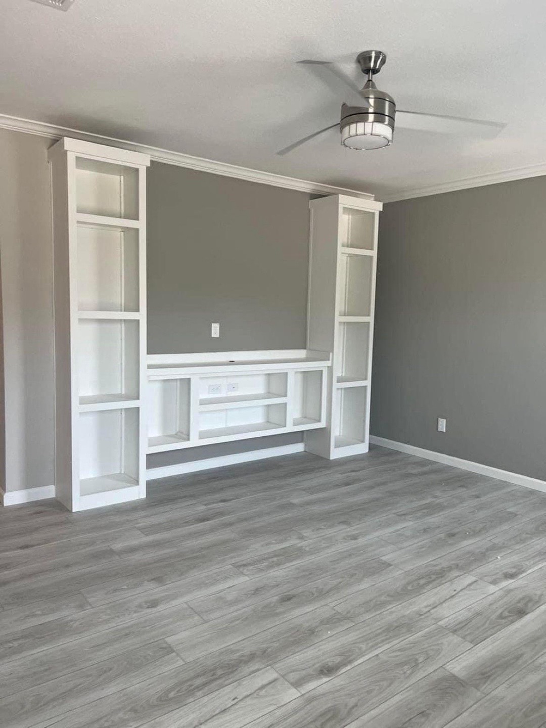 Empty room with a modern gray and white color scheme, featuring a built-in white entertainment center on a gray wall, light wood flooring, and a sleek ceiling fan.