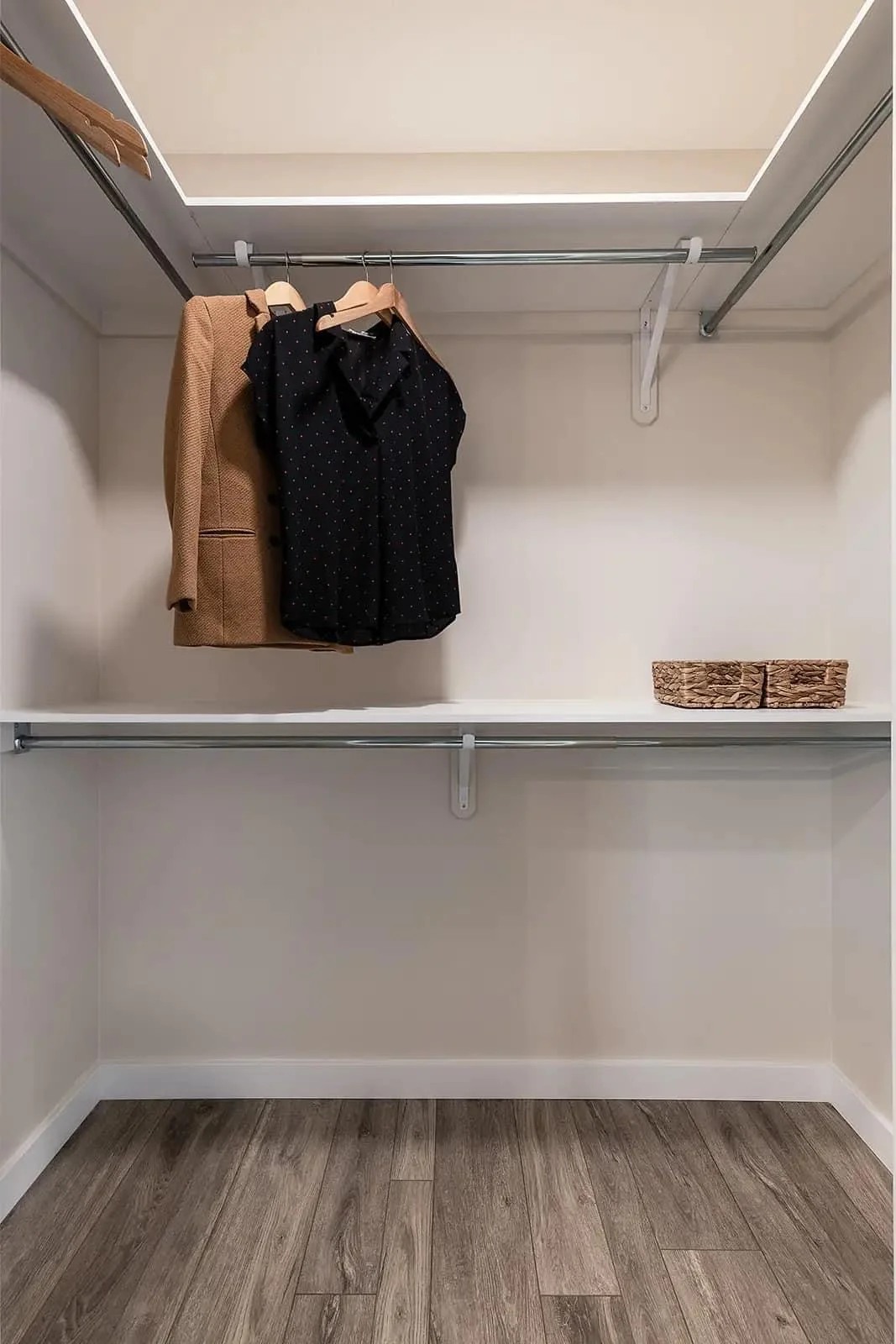 Spacious empty closet with light wood floor, white walls, and two shelves. A beige coat and black blouse hang neatly, with woven baskets nearby.