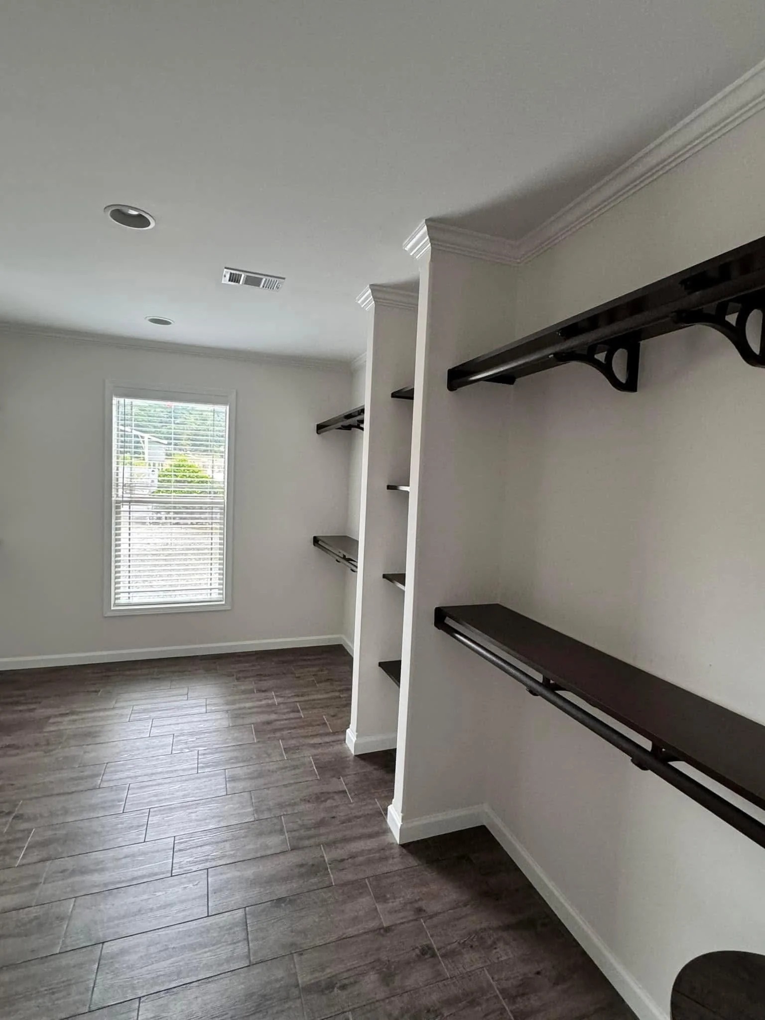 Spacious walk-in closet with dark wooden shelves and rods on white walls. Window on left lets in natural light, highlighting the dark tile floor.