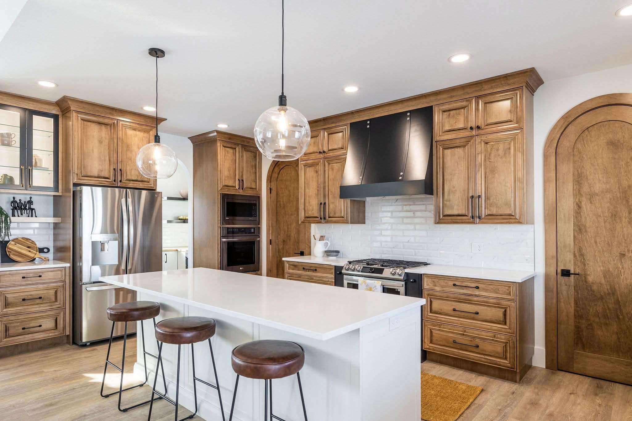 A modern kitchen with wooden cabinets, white countertops, and stainless steel appliances. Three leather bar stools line the central island, under warm pendant lights.
