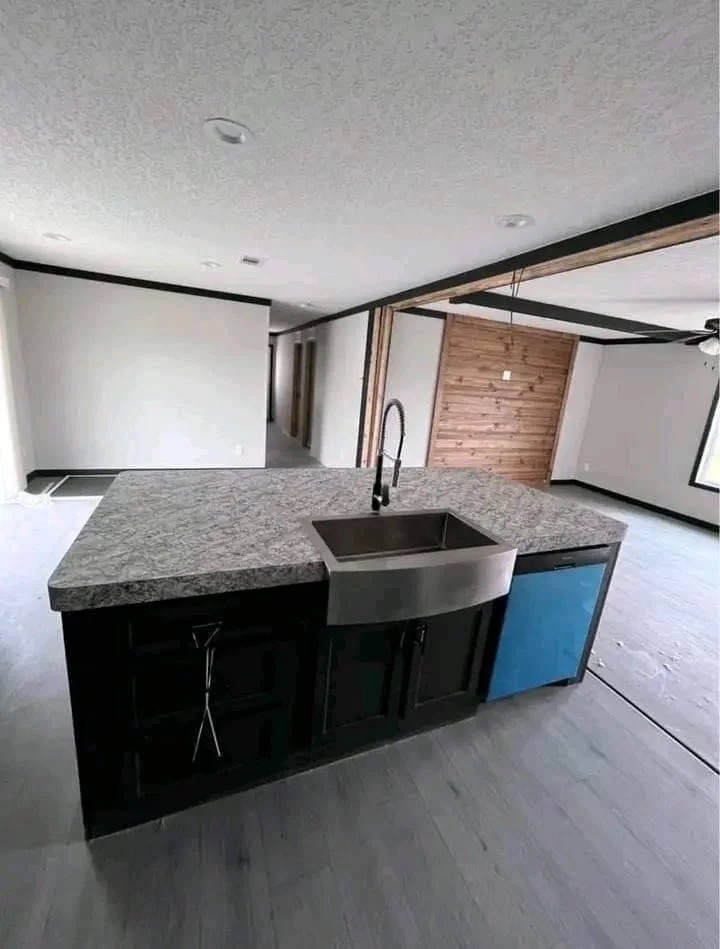 Modern kitchen with a granite island in the foreground, featuring a large stainless steel sink. The open layout includes wood accents and light flooring, evoking a sleek and inviting atmosphere.