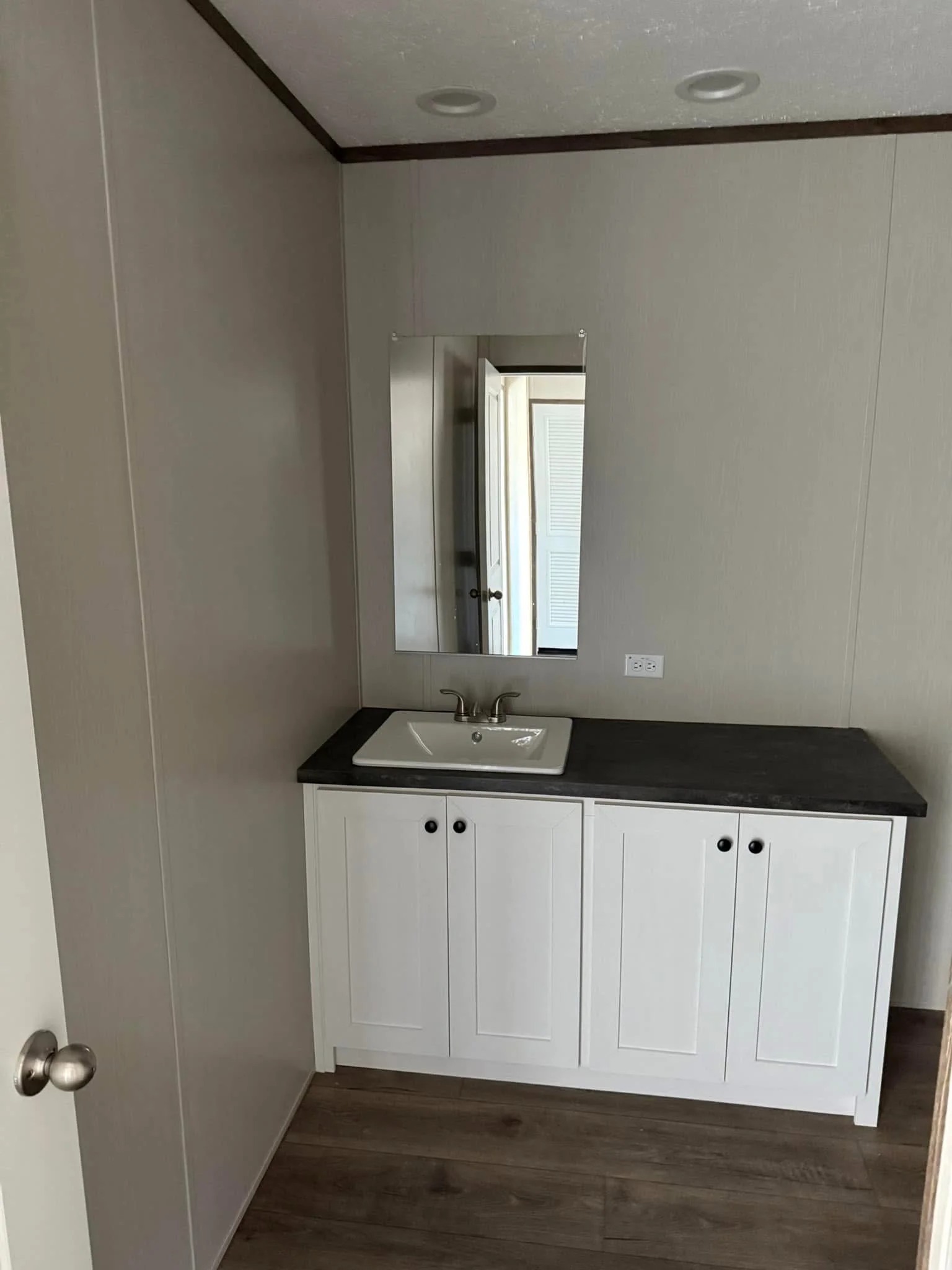 Simple bathroom with white cabinet, dark countertop, and rectangular mirror. Neutral walls, wood floor; minimalist and clean design.