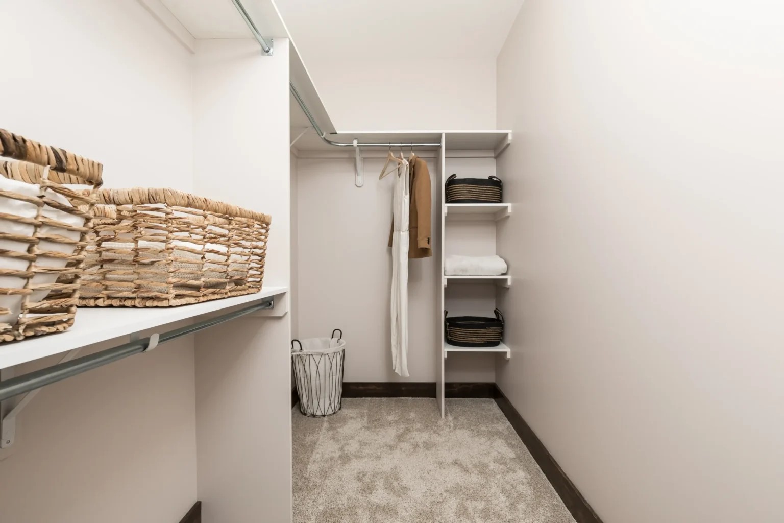 A minimalist walk-in closet with beige walls and carpet. It features wicker baskets on a shelf, hanging clothes, and neatly organized items on white shelves.