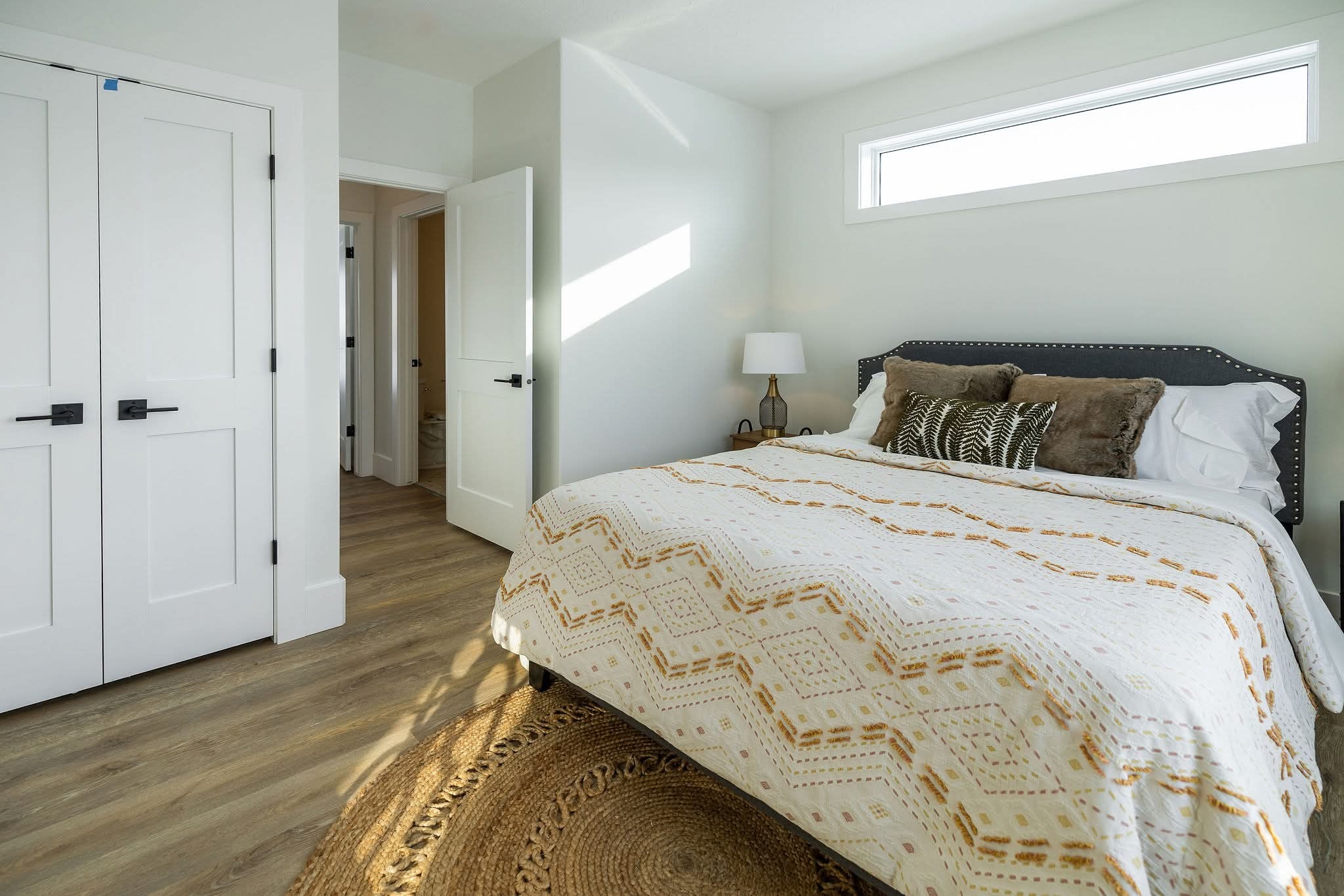 Modern bedroom with a cozy vibe features a queen bed, patterned quilt, and decorative pillows. Light streams through a high window, illuminating the space.