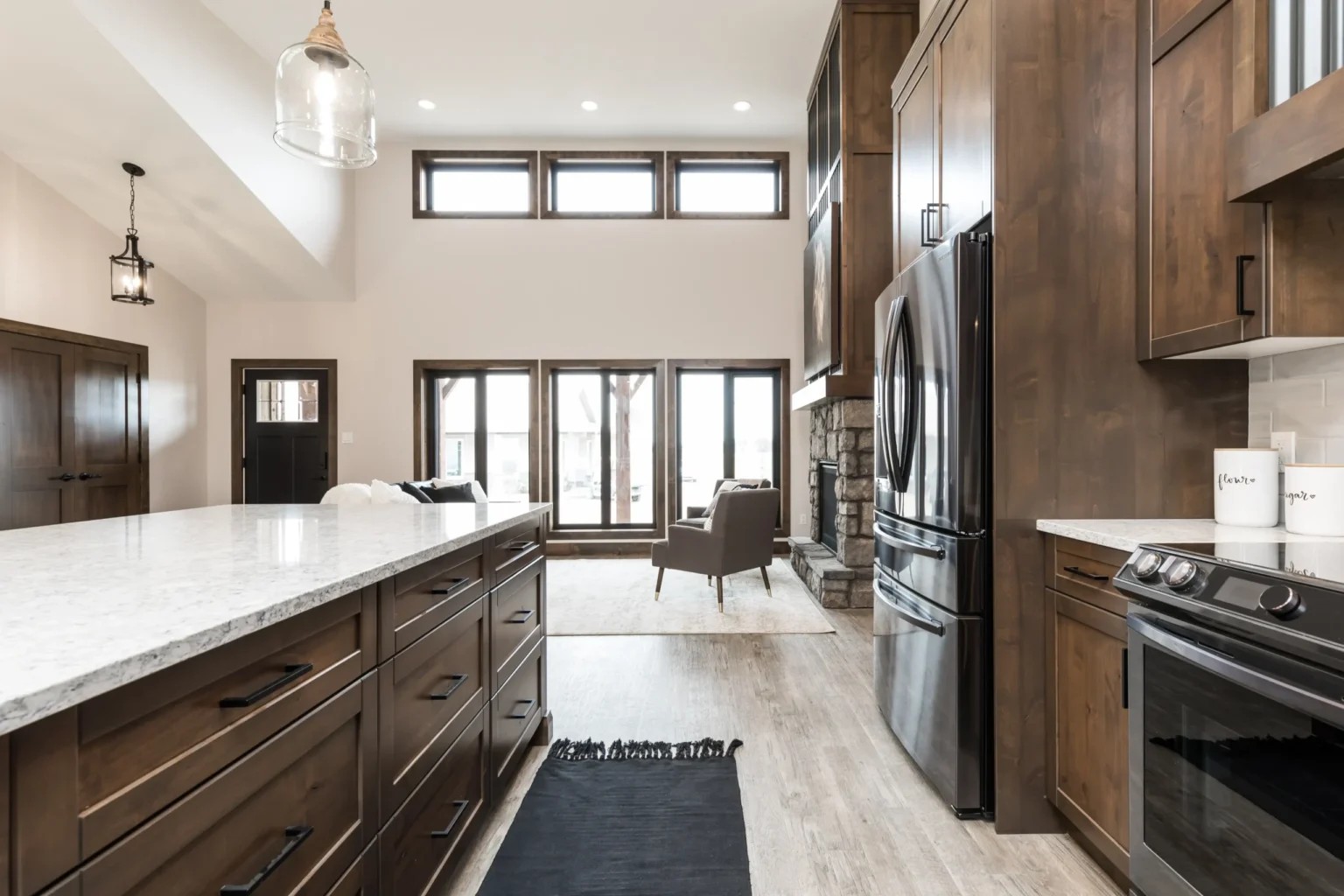 Spacious kitchen and living area with high ceilings, large windows, and wood cabinets. Features a marble island, black appliances, and modern decor.