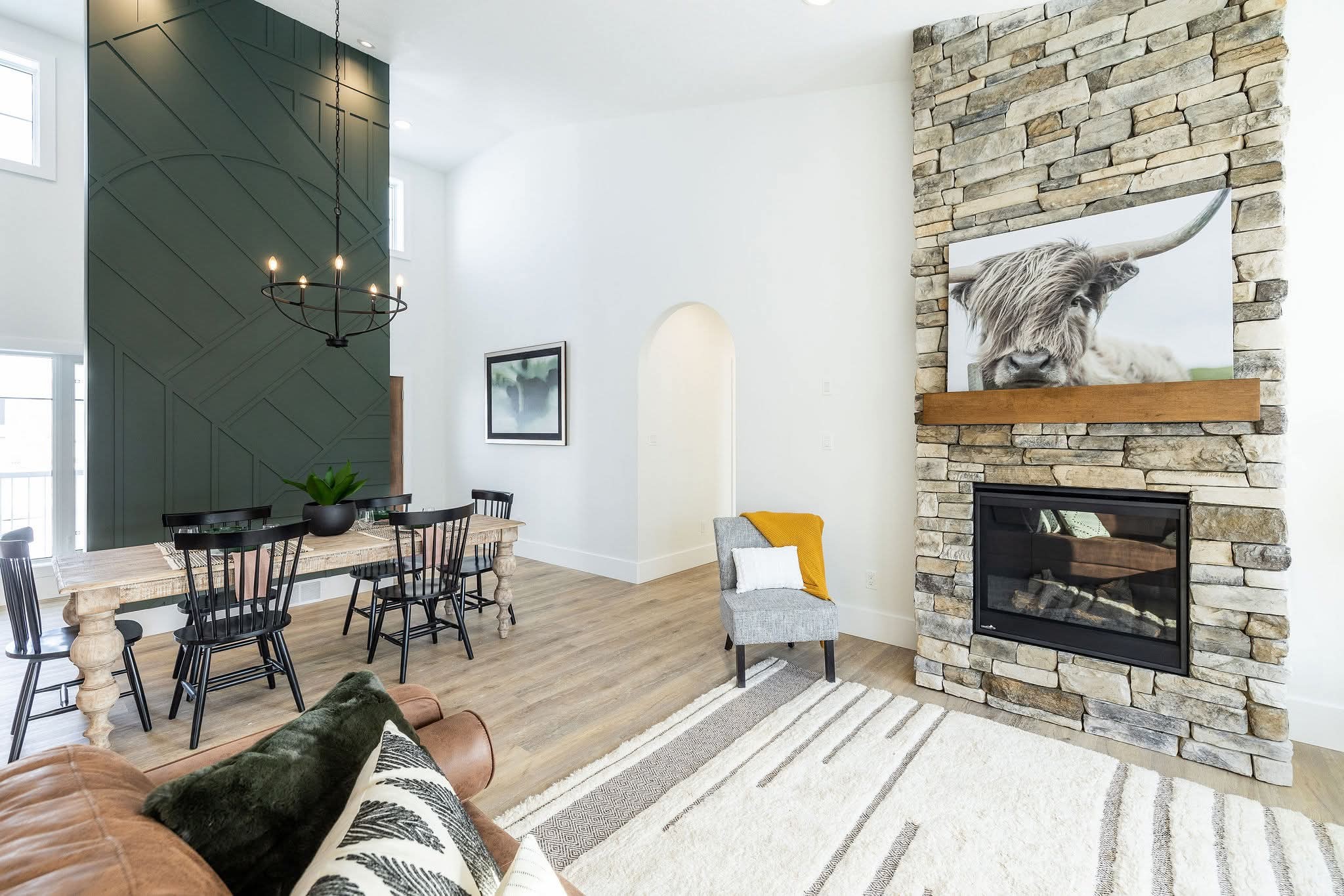 Modern living room with a stone fireplace featuring a cow portrait. Dark green accent wall, chandelier, and dining area create a cozy, stylish ambiance.