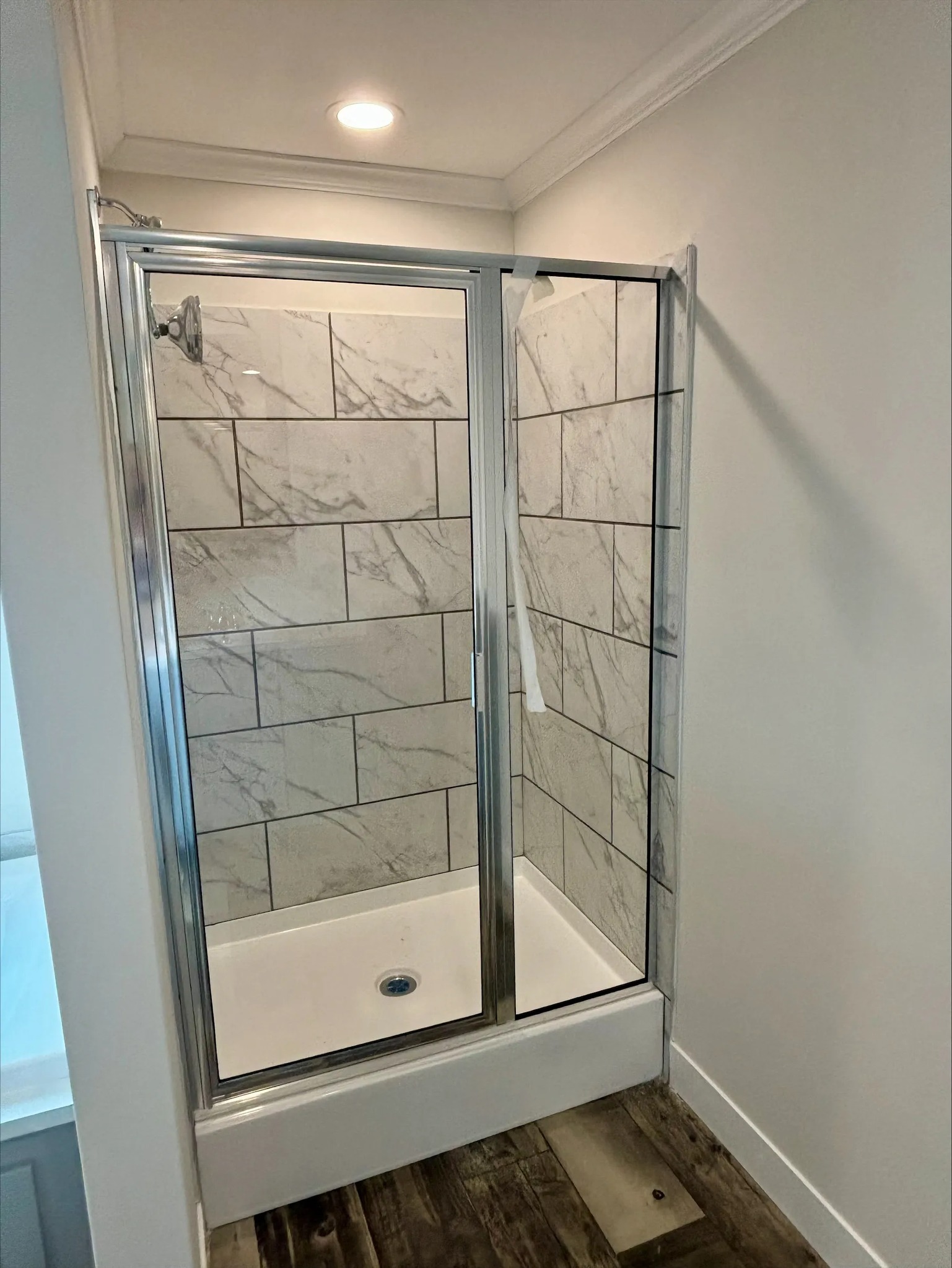 "Modern shower stall with sliding glass doors, marble-patterned tile walls, white shower base, and dark wood-look floor."