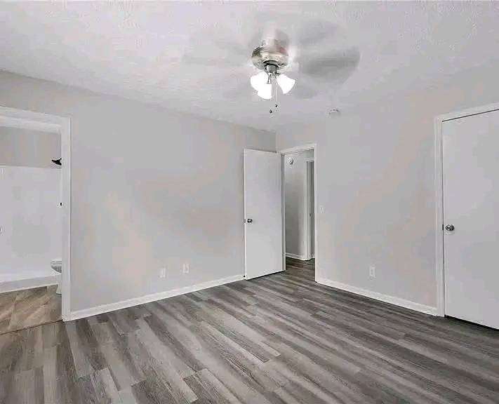 Empty room with light gray walls, wood floors, and a ceiling fan. Open doors lead to a bathroom and hallway, creating an inviting, serene space.
