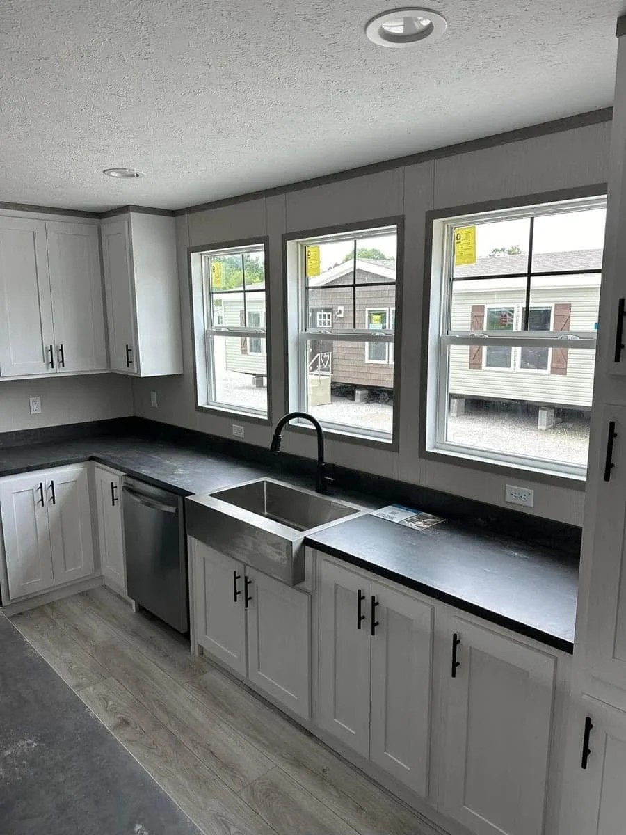 Modern kitchen with white cabinets, black countertops, and stainless steel sink. Sunlight from three windows creates a bright, open ambiance.