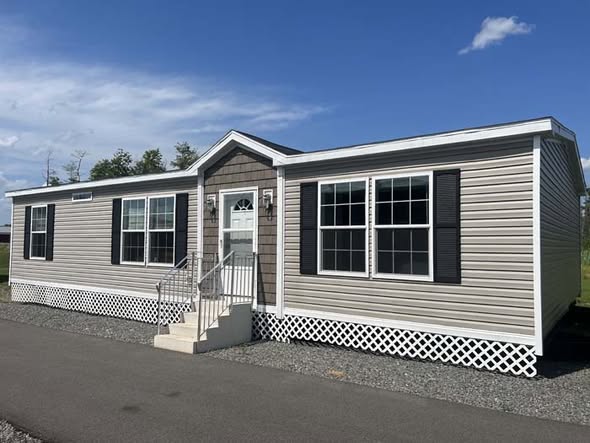 Modern grey manufactured home exterior with black window shutters, white lattice skirting, and a peaked roof entry.