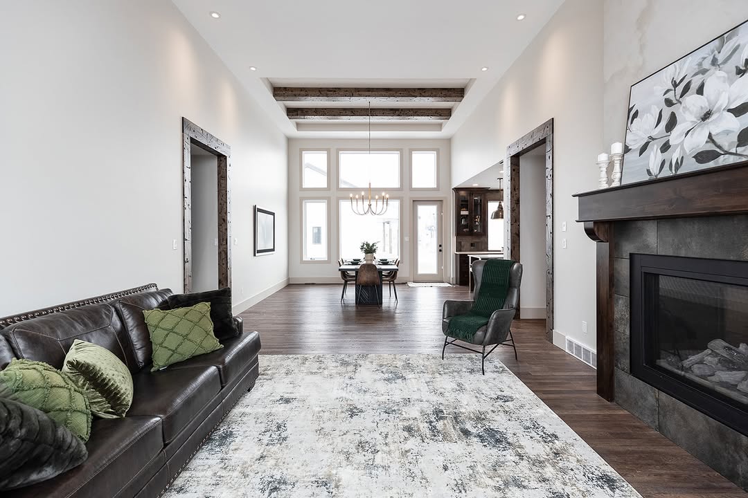 Spacious modern living room with wooden floors, dark leather sofa, green pillows, and a large rug. Large windows and a dining table lead to natural light. Cozy fireplace adds warmth.