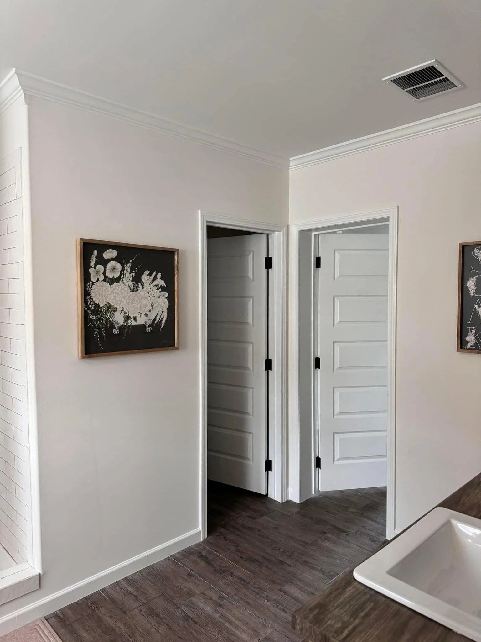 A minimalist bathroom corner with white walls, dark wood flooring, and a wooden vanity. Two white paneled doors are open. Floral artwork hangs on the wall.