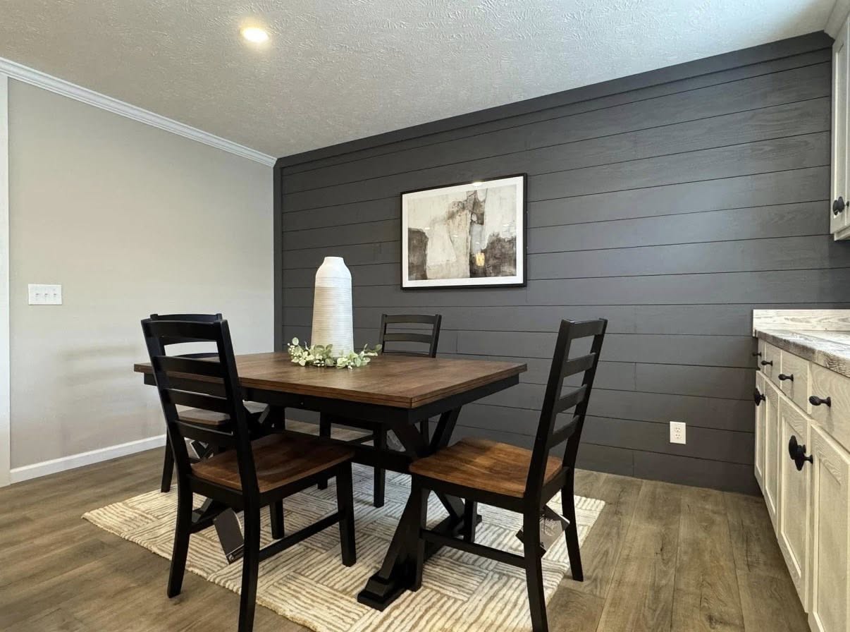 Dining area with a wooden table, four black chairs, and a textured vase centerpiece. Dark shiplap wall with abstract art, minimalist and modern feel.