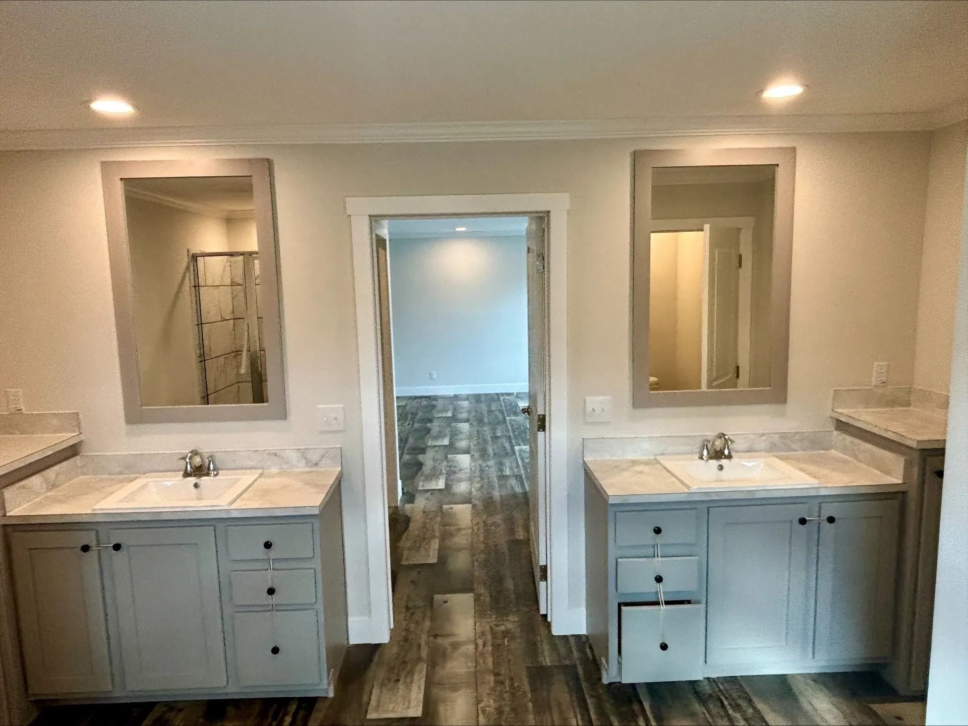 A collection of real estate images showcasing multiple modern farmhouse-style manufactured homes with high-end interior and exterior finishes. The exterior views display various designs, including a light blue home with a large gabled front and vaulted windows, and a navy blue home featuring white shutters and a covered porch with stone-accented pillars. Inside, the homes offer open-concept living areas with vaulted ceilings, exposed dark wood beams, and prominent floor-to-ceiling stone fireplaces. Kitchens are designed with a mix of white and grey cabinetry, large islands with marbled or dark countertops, farmhouse sinks, and industrial-style chandeliers with Edison bulbs. Dining areas feature rustic wood tables, forest-themed wall art, and large windows. The bathrooms highlight luxurious amenities such as freestanding soaking tubs, double vanities with marbled tops, and walk-in showers with marble-patterned tile and glass partitions. Design details throughout include light and dark wood-plank flooring, sliding barn doors, and contemporary neutral decor.