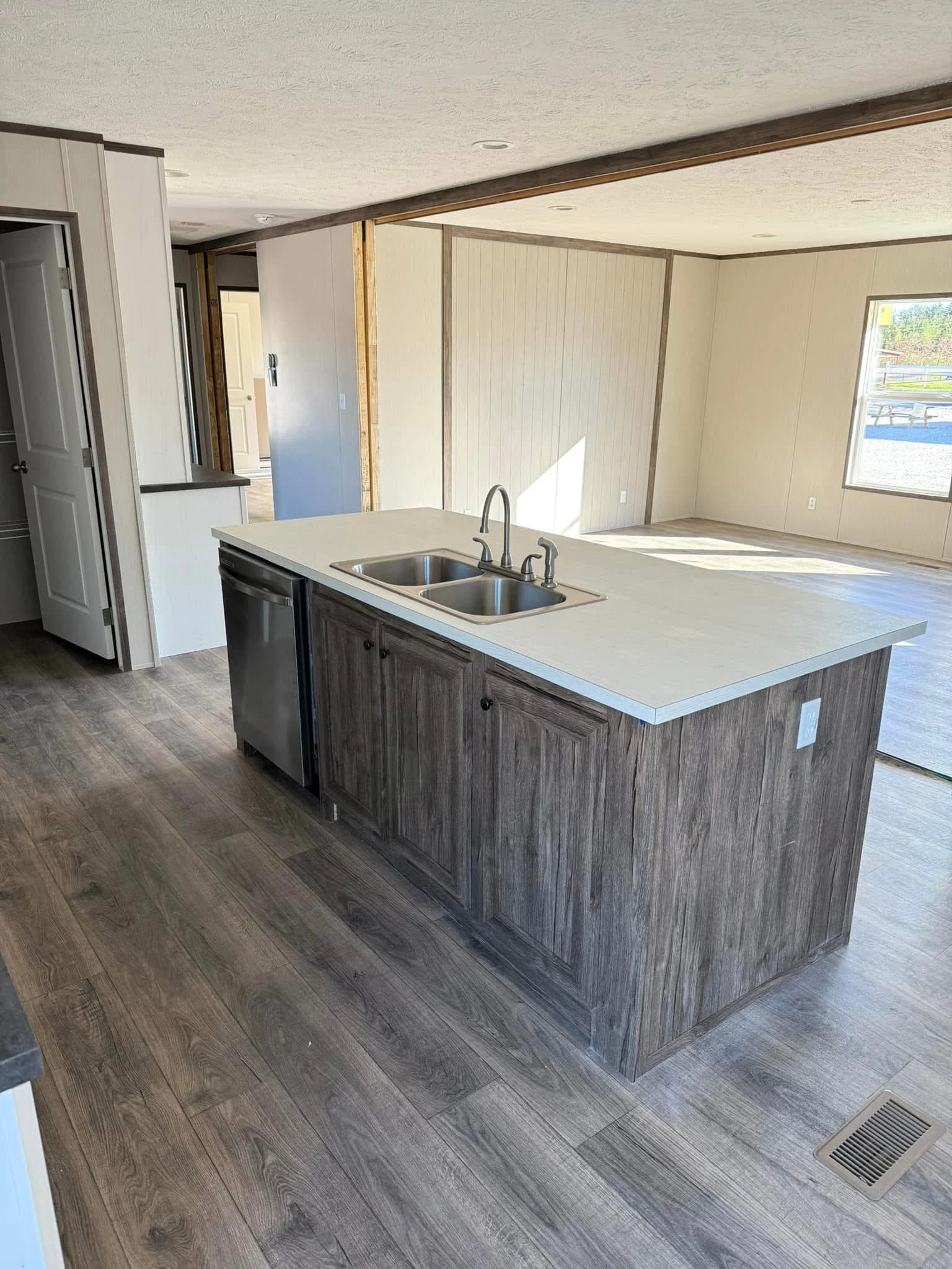 Modern kitchen interior with sleek wood flooring and a central island featuring a double sink. Bright, airy feel with natural light from a window.
