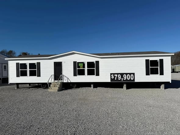 A white prefabricated home sits on a gravel lot under a clear blue sky. Black shutters frame the windows, and a $79,900 price is displayed prominently.