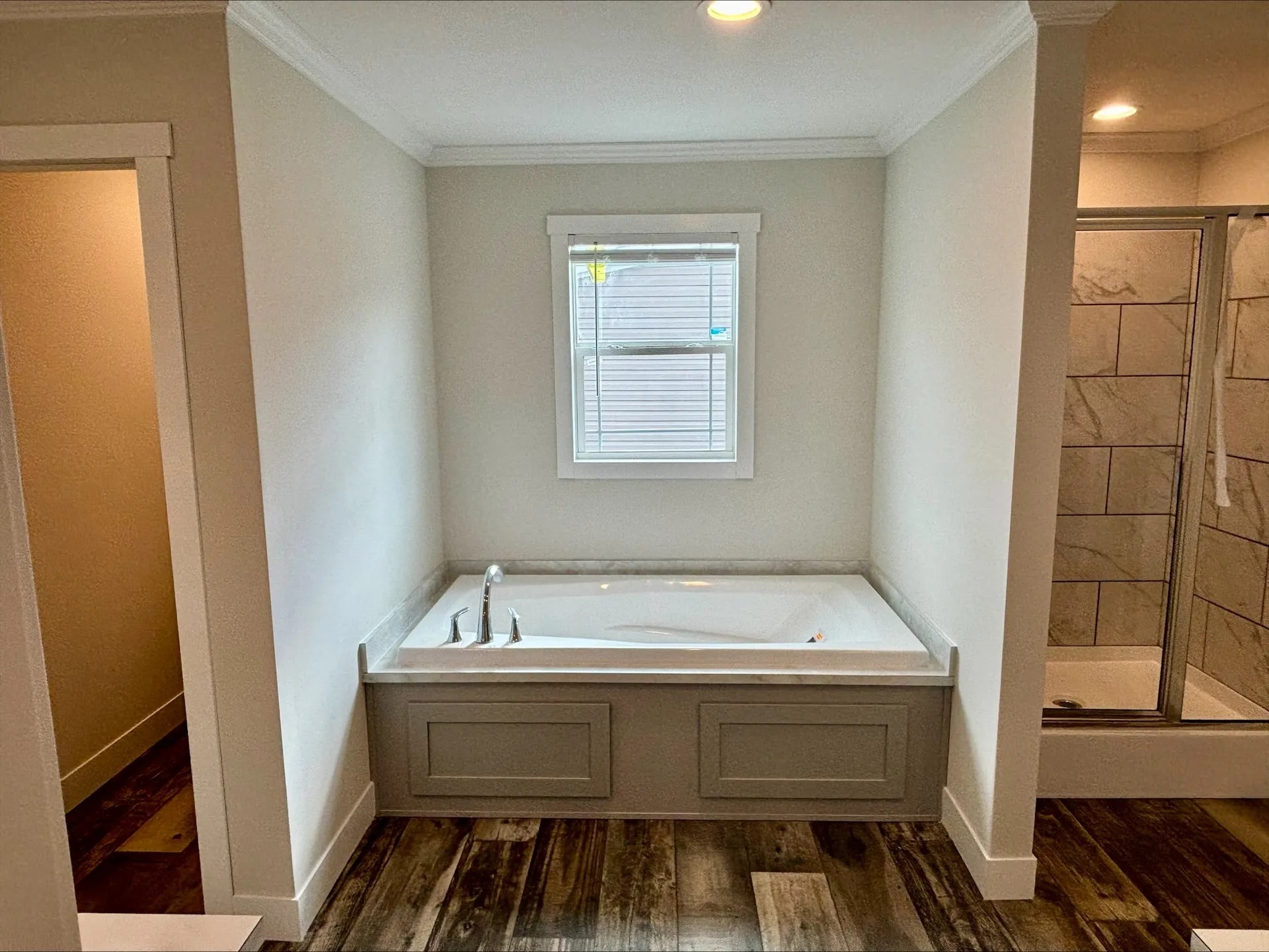 "Bathroom with a built-in rectangular bathtub under a small window, light gray paneled front, chrome faucet, beige walls, dark wood-look flooring, and a tiled glass shower enclosure visible to the right."
