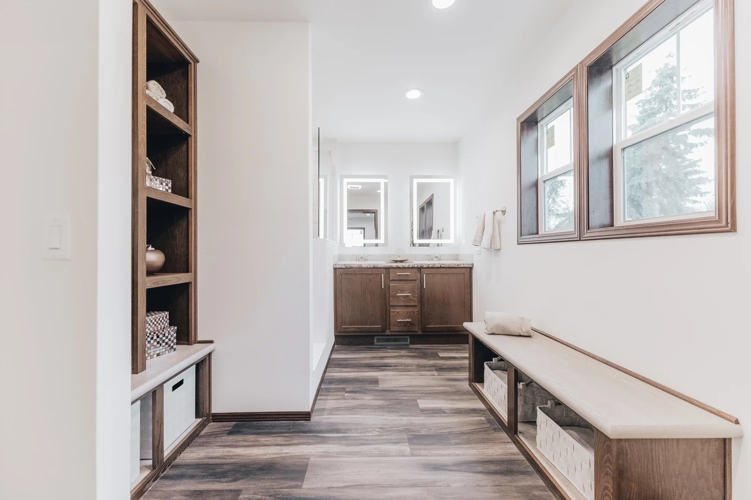 Minimalist hallway with wooden floors, built-in bench and shelves, leading to a bathroom with wooden cabinets and two illuminated mirrors. Bright and tidy ambiance.