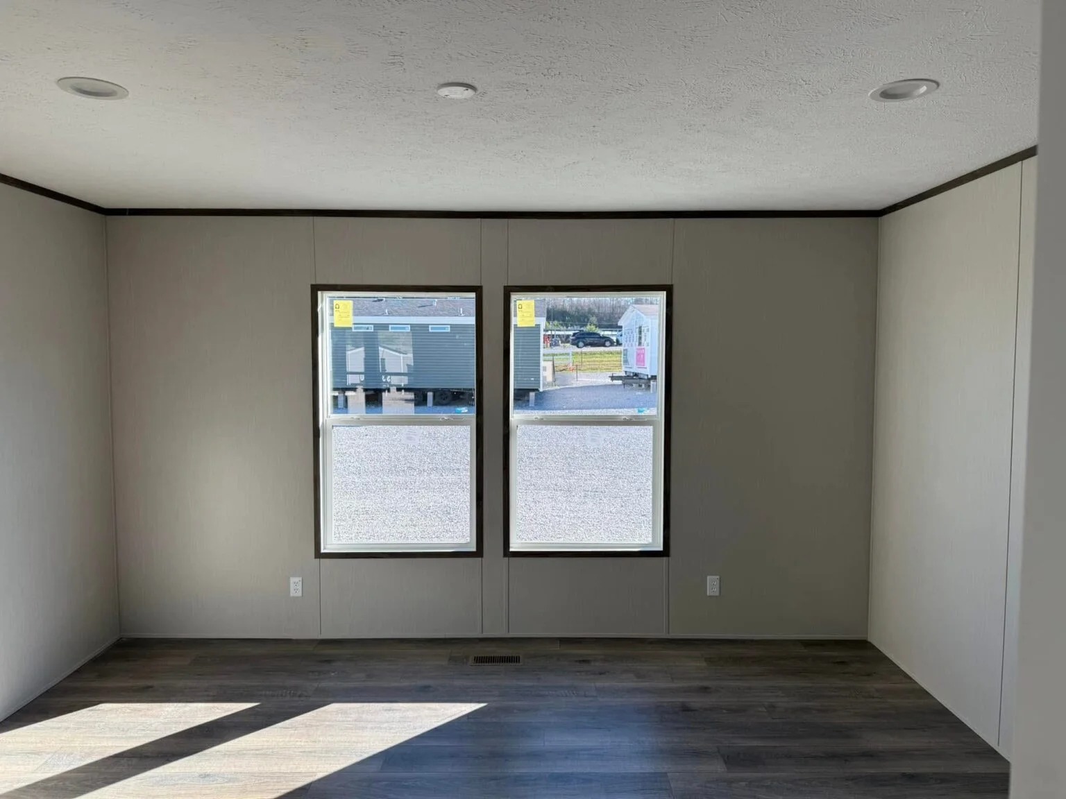 Empty room with beige walls and dark wood flooring, featuring two large windows showing a parking lot with trailers. Sunlight casts shadows on the floor.