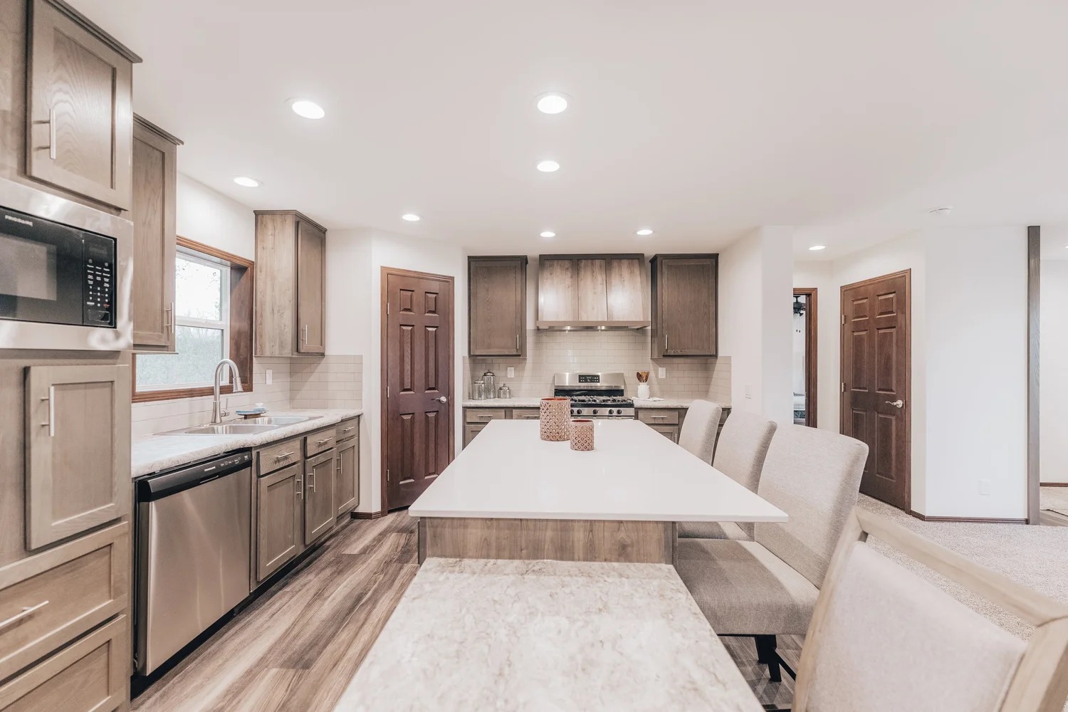 Modern kitchen with wooden cabinets and stainless steel appliances. Features a large island with beige bar chairs, wood flooring, and recessed lighting. Cozy and inviting.