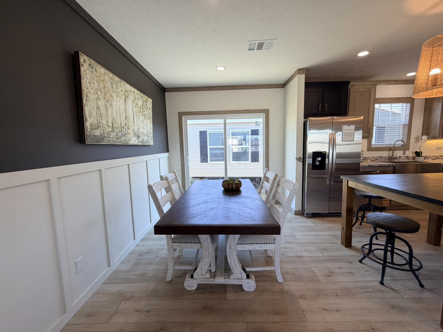 Bright dining area with a long wooden table and six chairs, light wood flooring, dark accent wall, art piece, and modern kitchen in the background.