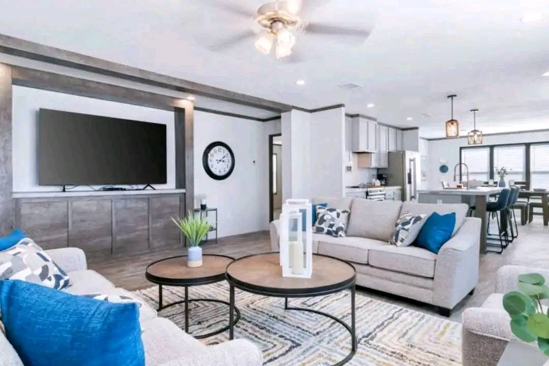 Modern living room with neutral tones features two light gray sofas accented with blue and patterned pillows. A round coffee table stands on a textured rug. Large flat-screen TV is mounted on the wood-paneled wall. The open-concept kitchen in the background includes stainless steel appliances, pendant lighting, and a dining area. The space feels airy and inviting.