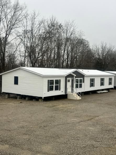 Comprehensive luxury manufactured home showcase featuring expansive double-wide floor plans, including the white board-and-batten APH 1662C and dark charcoal models with rustic wood shutters. High-end interior highlights include open-concept living rooms with integrated electric fireplaces and shiplap accent walls, gourmet kitchens with large marble-look waterfall islands and herringbone tile backsplashes, and custom laundry rooms with marble-look countertops. The design is elevated by vaulted ceilings with dark wood beams, master suites featuring black accent walls and cowhide rugs, luxury bathrooms with freestanding soaking tubs and LED-mirrored double vanities, and a peaceful covered front porch with a dark wood-plank ceiling and a hanging wicker egg chair.