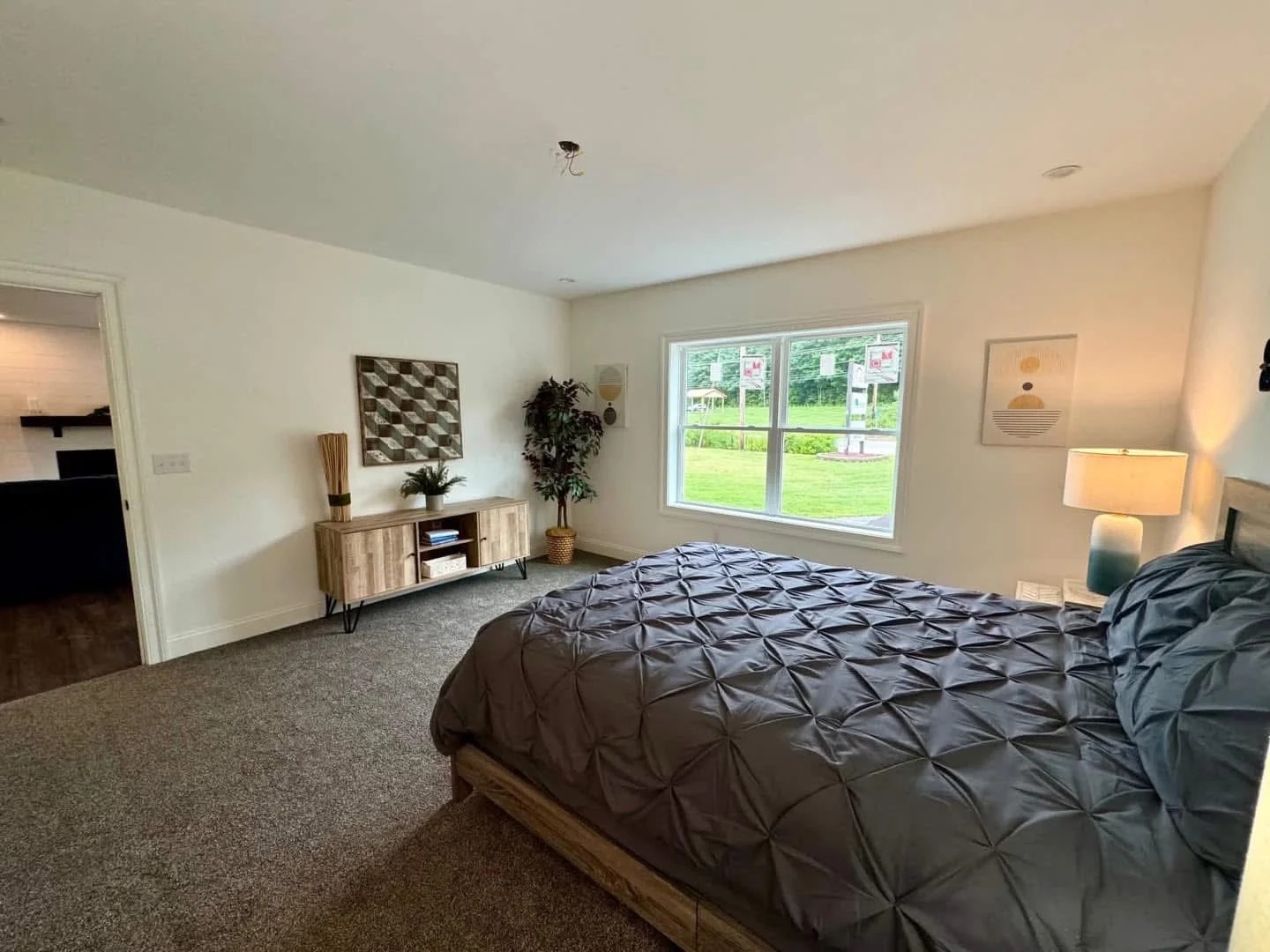 A spacious bedroom with a large bed covered in a textured gray comforter. A wide window offers a view of greenery, while minimal decor creates a calm, modern ambiance.