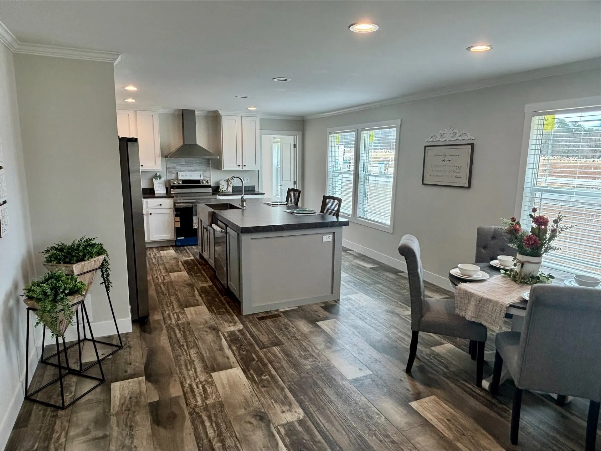 A comprehensive real estate gallery showcasing several modern farmhouse-style manufactured homes. Exterior views include a light blue home with a prominent gabled front and large vaulted windows, as well as a dark charcoal home with white trim and a covered porch. Interior living spaces feature open-concept layouts with vaulted ceilings, exposed dark wood beams, and floor-to-ceiling stone fireplaces. Kitchens are designed with a mix of white and grey cabinetry, large islands with marbled or dark countertops, farmhouse sinks, and industrial lighting. Dining areas feature rustic wooden tables, forest-themed wall art, and ample natural light from large windows. Bathrooms include high-end finishes like freestanding soaking tubs, double vanities with marbled tops, and walk-in showers with marble-patterned tile and glass partitions. Design details throughout include light wood-plank flooring, sliding barn doors, and contemporary decor.