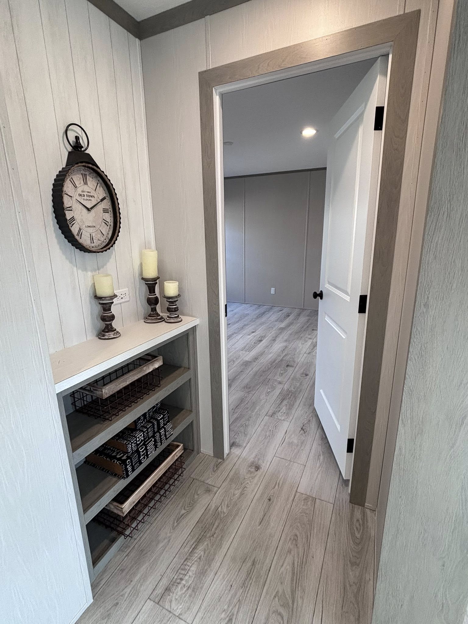 A comprehensive look at a modern farmhouse-style interior featuring a neutral grey and white color palette with light wood-plank flooring. The dining area is furnished with a dark wood-top table, white chairs, and a matching bench, accented by a large black-and-white forest canvas and rustic wall decor. The kitchen boasts a large island with a swirling grey marbled countertop, cushioned barstools, and an industrial-style chandelier with Edison bulbs, complemented by grey cabinetry, a farmhouse sink, and a sliding wooden barn door. The bathroom includes a double vanity with marbled tops, a white freestanding soaking tub, and a walk-in shower featuring marble-patterned tiles and a glass partition. An entryway view shows a built-in shelving unit with decorative candles and a vintage-style wall clock leading into a bright, open room.
