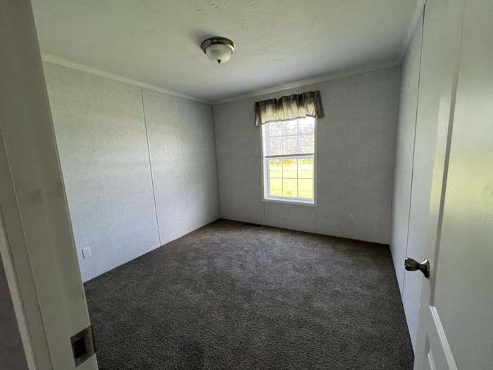 Bright and airy manufactured home bedroom with neutral walls, grey carpeting, and a large window overlooking the lot.