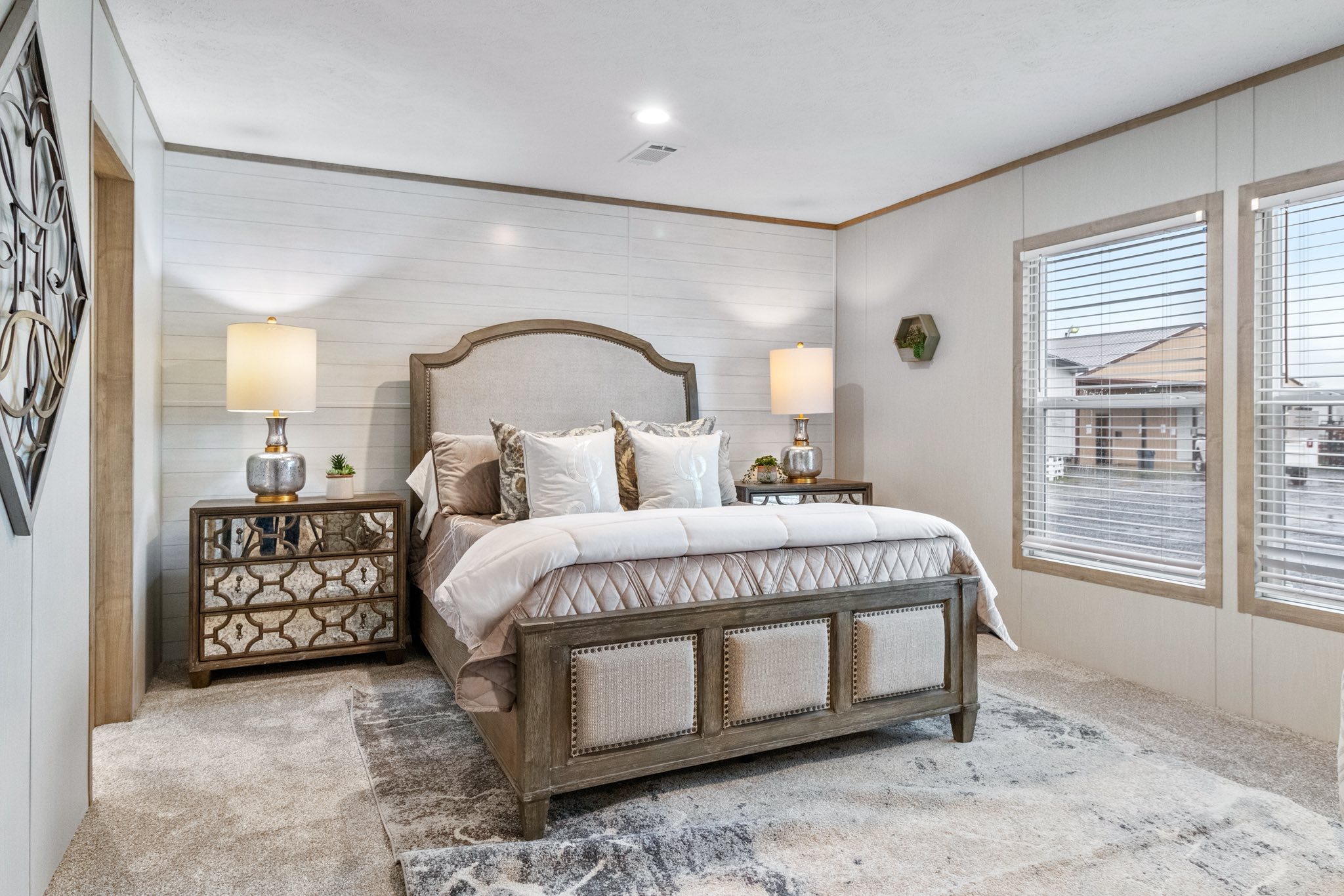 Elegant bedroom with a plush beige bed, mirrored nightstands, warm lamps, and large windows. The room is cozy, inviting, and well-lit. Neutral tones dominate.