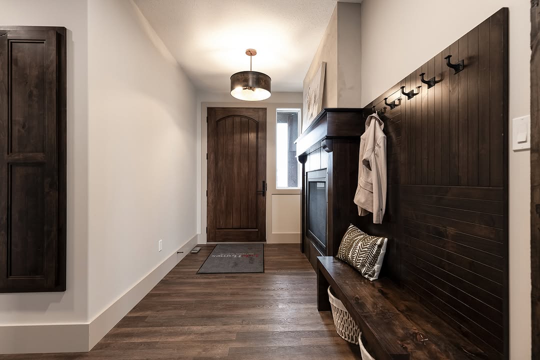 Cozy entryway with wooden bench and coat hooks on the right, wooden door and a narrow window ahead, warm light fixture overhead, welcoming ambiance.