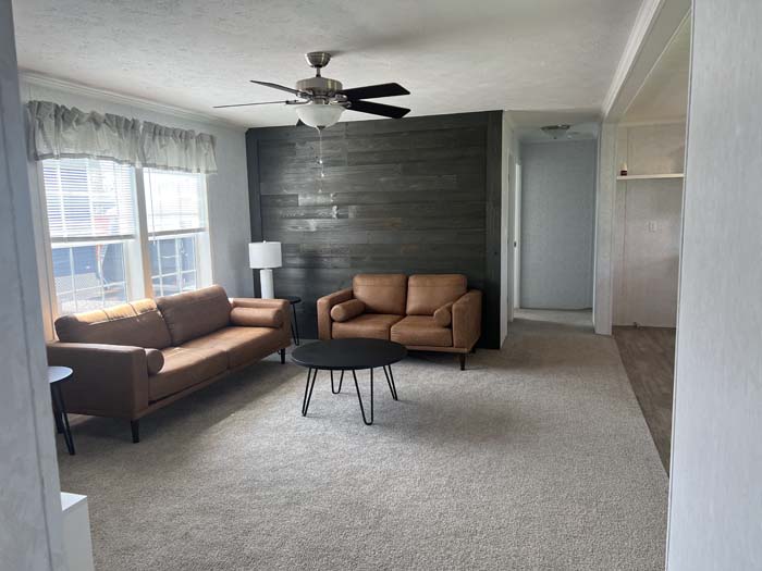 Spacious manufactured home living room featuring a dark wood accent wall, tan leather sofas, a black coffee table, and plush grey carpeting.