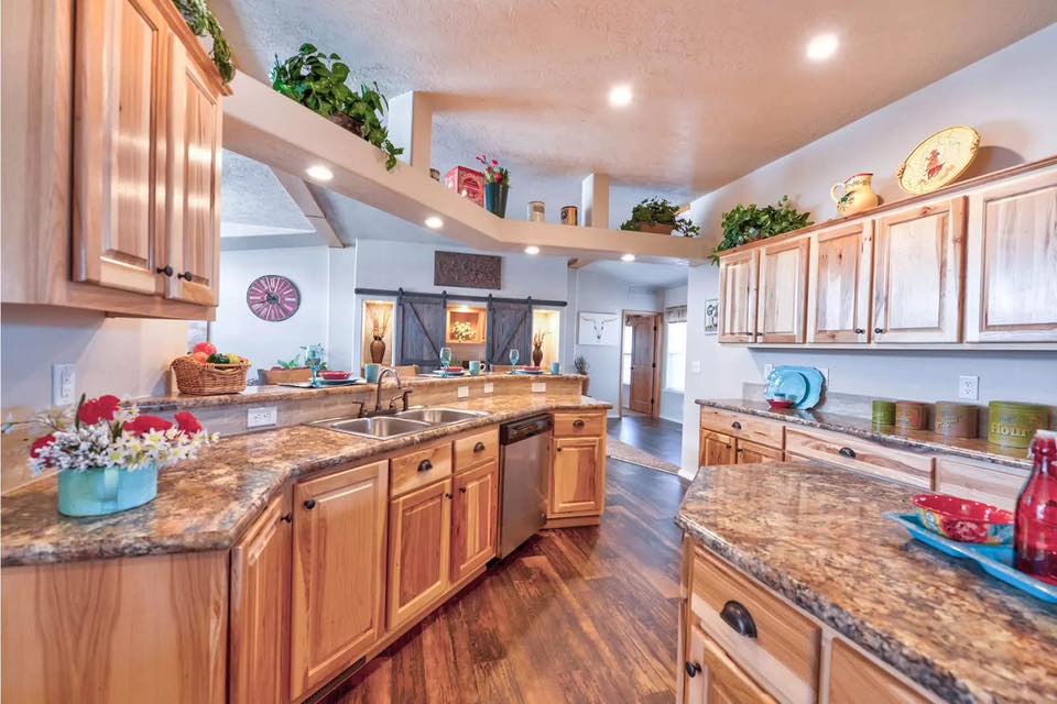 Modern rustic kitchen interior featuring natural wood cabinetry, granite countertops, and a double stainless steel sink with decorative indoor plants and warm recessed lighting.