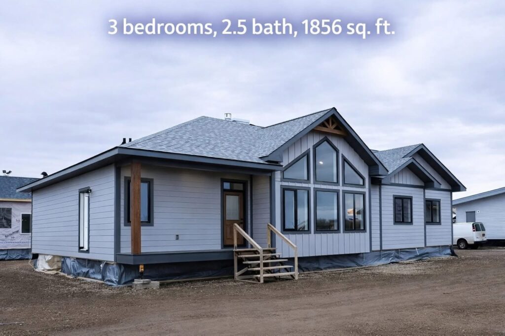 A comprehensive gallery showcasing a modern farmhouse-style home with light wood-plank flooring and a neutral grey and white color palette. The exterior image displays a light blue manufactured home with a prominent gabled front featuring large windows and a wooden porch. The interior dining area includes a dark wood-top table with white legs, matching chairs, a bench, and a large black-and-white forest canvas. The kitchen features a large island with a swirling grey marbled countertop, white cushioned barstools, and an industrial-style black chandelier, complemented by grey cabinetry, a farmhouse sink, and a rustic wooden sliding barn door. Bathroom areas highlight a double vanity with marbled tops, a white freestanding soaking tub, and a walk-in shower with marble-patterned tiles and a glass partition. An entryway view shows a built-in shelving unit with decorative candles and a vintage-style wall clock leading into a bright, open room.