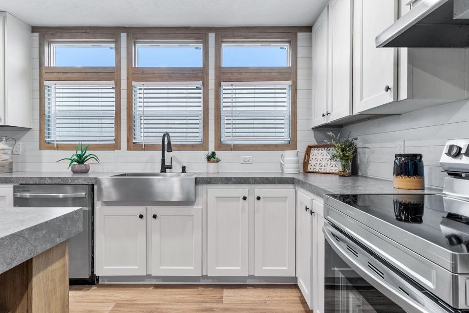 Modern kitchen with white cabinets, a stainless steel sink, and gray countertops. Three large windows with blinds provide natural light. Cozy and clean ambience.