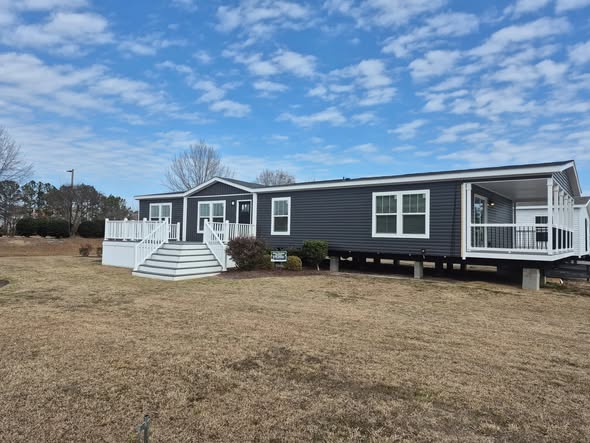 A modern gray mobile home sits on a spacious grassy lot under a partly cloudy sky. The home features white railings and steps, conveying a welcoming feel.