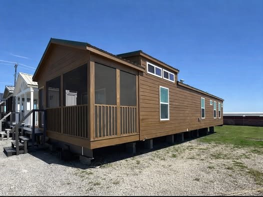 A modern tiny house with a wooden exterior and screened porch sits on a gravel lot under a clear blue sky. It is elevated on blocks with a few windows visible.