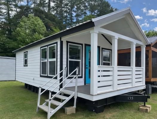 This extensive image gallery showcases a variety of luxury tiny homes, park models, and manufactured homes from the "Boujee XL" series, highlighting a refined modern farmhouse aesthetic through both exterior and interior designs. The exteriors feature a mix of white and cream board-and-batten siding accented by charcoal gables, gabled front porches with black or white pillars, and sleek metal roofing in white or dark grey. The interiors emphasize open-concept living with vaulted ceilings, exposed wood-look beams, and large transom windows that provide abundant natural light to spaces finished with wide-plank wood-look flooring and crisp white shiplap or neutral walls. Gourmet kitchens are equipped with stainless steel appliances, farmhouse sinks, and cabinetry ranging from light oak to elegant white, often featuring built-in breakfast nooks or large islands with distressed wood-look countertops. Functional luxury is further integrated through features such as lofted sleeping areas with black metal railings and wooden ladders, industrial-style ceiling fans, carpeted bedrooms with transom windows, and modern bathrooms with marble-patterned walk-in tile showers.