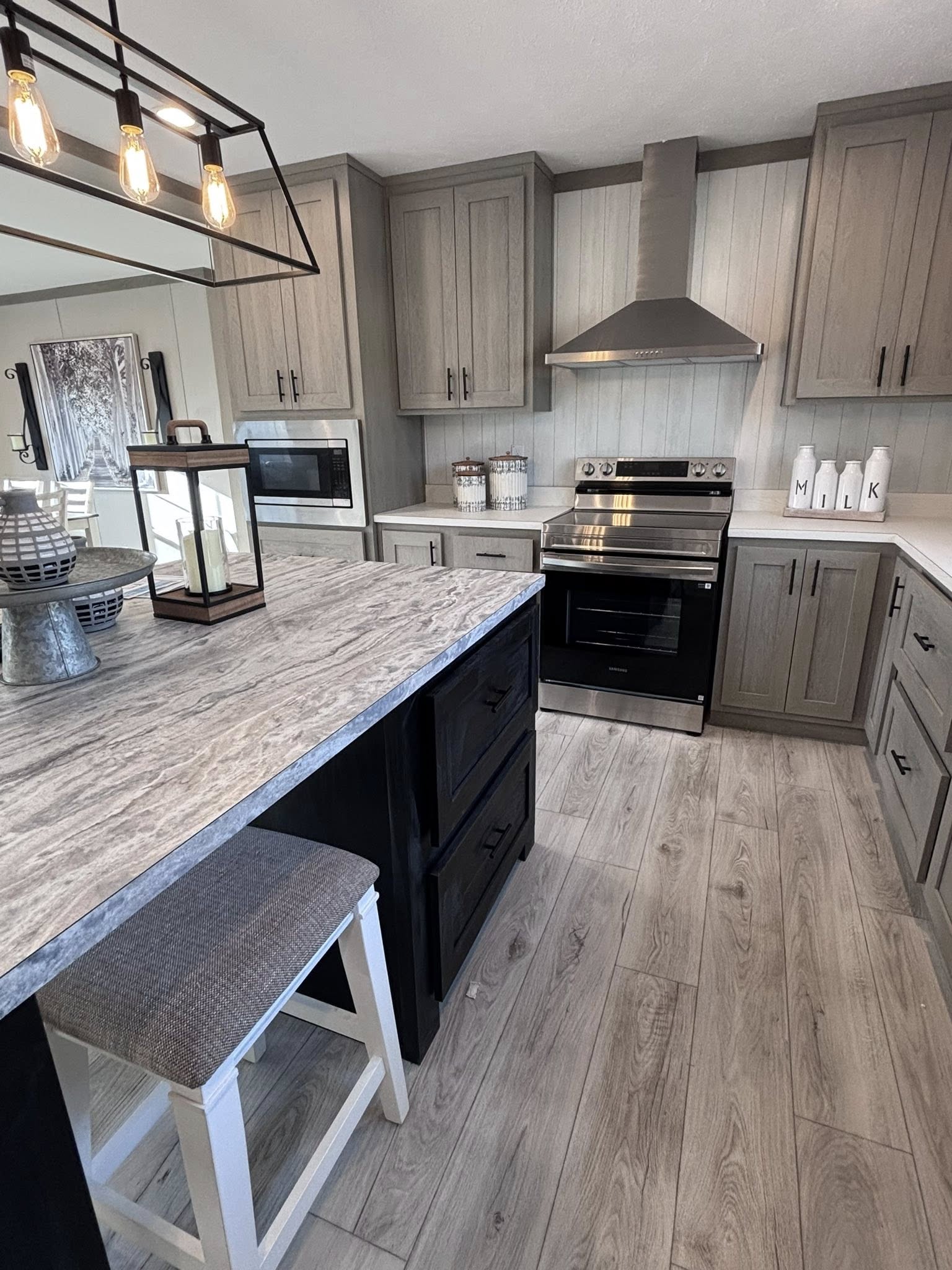 A collage of images showcasing a modern farmhouse interior with a grey and wood-tone color palette. The dining area features a dark wood-top table with white legs, matching chairs, a bench, and rustic wall decor including a large forest path canvas. The kitchen contains a large island with a swirling grey marbled countertop, cushioned white barstools, and an industrial-style chandelier with Edison bulbs. Additional kitchen details include a white farmhouse sink, grey cabinetry, stainless steel appliances, and a wooden sliding barn door leading to a pantry.