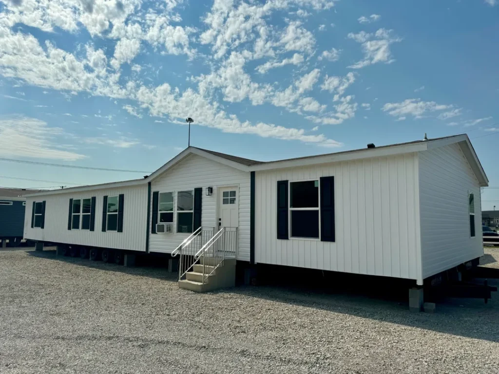 A white, single-story modular home with black shutters sits on a gravel lot under a blue sky with wispy clouds. Stairs lead to a central door.