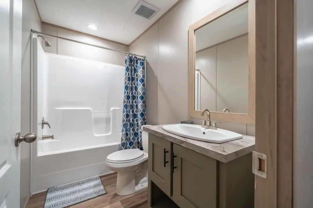 Modern bathroom with beige walls, a white bathtub with blue patterned shower curtain, and a sink on a green vanity. Wood-like flooring and calm ambiance.