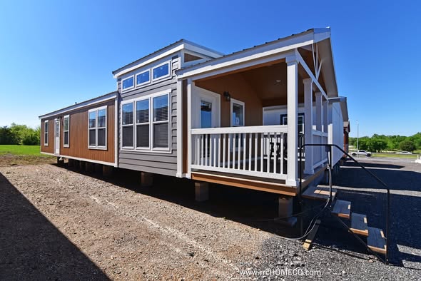 A modern manufactured home with tan and white siding under a clear blue sky. It features large windows, a small porch with a railing, and steps leading up.