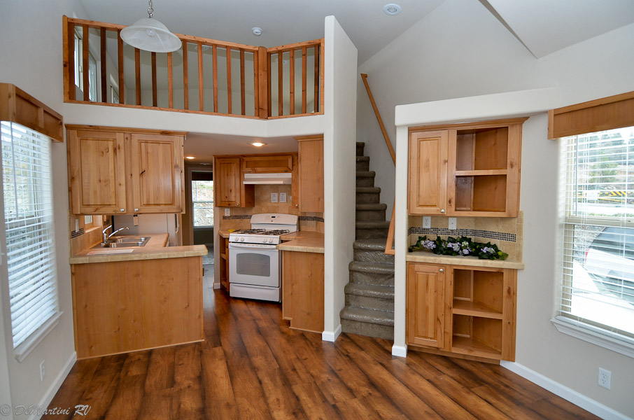 Classic white and soft sage green board-and-batten tiny homes featuring grand gabled front porches with black metal railings and sleek metal roofing.

A striking premium park model with a dark charcoal exterior, a vibrant copper-tone metal roof, natural cedar wood pillars, and a massive wrap-around elevated wooden deck.

Traditional double-wide manufactured home with tan vinyl siding, decorative burgundy shutters, and a full-length pillared front porch on a gravel lot.

Rustic log-cabin style park model with warm wood siding, a green metal roof, and an integrated porch with a decorative wreath.

Interior Living & Custom Design:

Modern open-concept living areas featuring vaulted ceilings with rustic dark wood beams, white shiplap walls, and integrated loft sleeping quarters with black metal or natural wood balustrades.

A cozy rustic cabin interior defined by all-over natural wood-planked walls and ceilings, featuring a dark grey sofa, a matching coffee table, and a large black industrial ceiling fan.

Traditional living space with neutral carpeting, patterned armchairs, and floor-to-ceiling drapery in a modern geometric print.

Gourmet Kitchens & Dining:

High-end kitchens featuring oversized islands and L-shaped layouts with charcoal or deep navy blue cabinetry, marble-look waterfall countertops, and professional-grade stainless steel appliances.

Rustic tiny home kitchen with natural wood cabinetry, black marble-look countertops, and a large stainless steel refrigerator.

Modern dining area with a circular dark wood table, a gold ring-style chandelier, and rustic wood-look flooring.

Spa-Inspired Bathrooms & Bedrooms:

Master bathroom featuring a walk-in tile shower with marble-look surrounds and sliding glass doors, paired with a grey vanity and rustic wood-look countertops.

Minimalist master bedroom with plush neutral carpeting, large windows for natural light, and a double-door closet system.