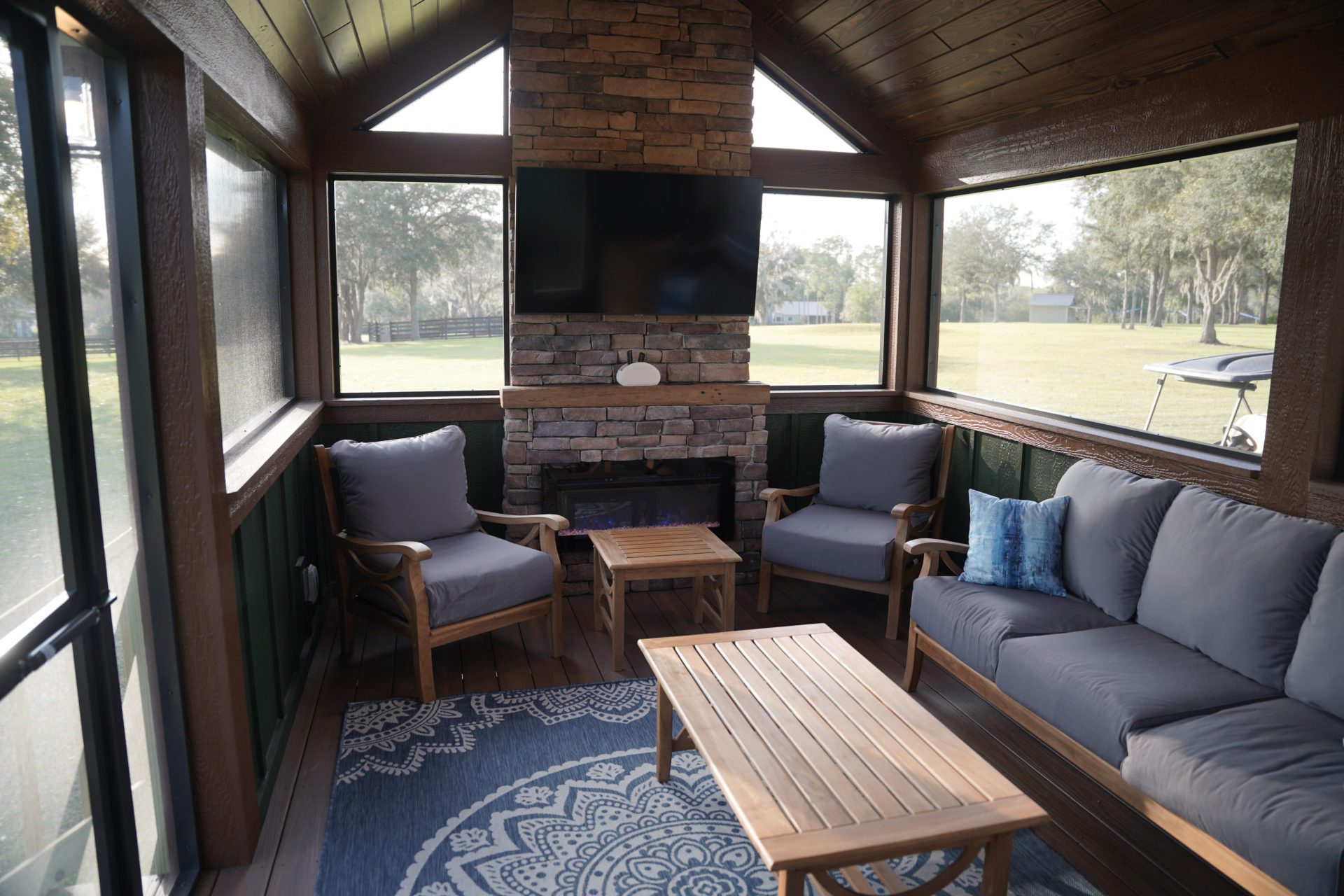 Cozy sunroom with large windows, featuring a stone fireplace, mounted TV, blue-gray sofas, wooden coffee table, and a patterned blue rug. Relaxing ambiance.