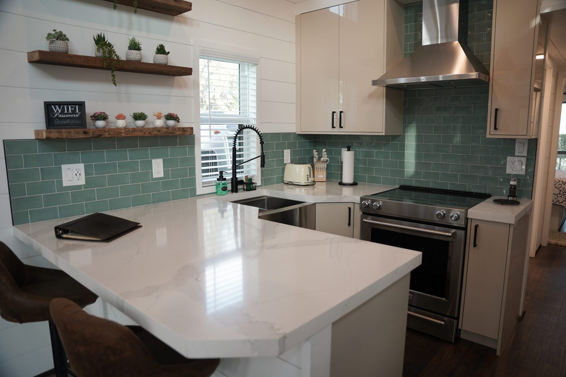 Modern kitchen with light green tile backsplash, white cabinets, and marble countertops. Features stainless steel appliances and wooden shelves with small plants. Cozy and clean ambiance.