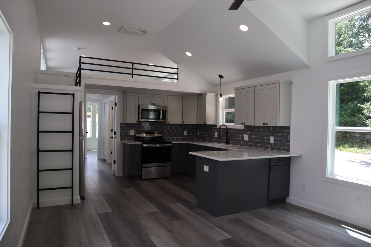 Modern kitchen with high ceilings, gray cabinets, and black backsplash. Features stainless steel appliances, a ladder to a loft, and large windows. Bright and airy space.