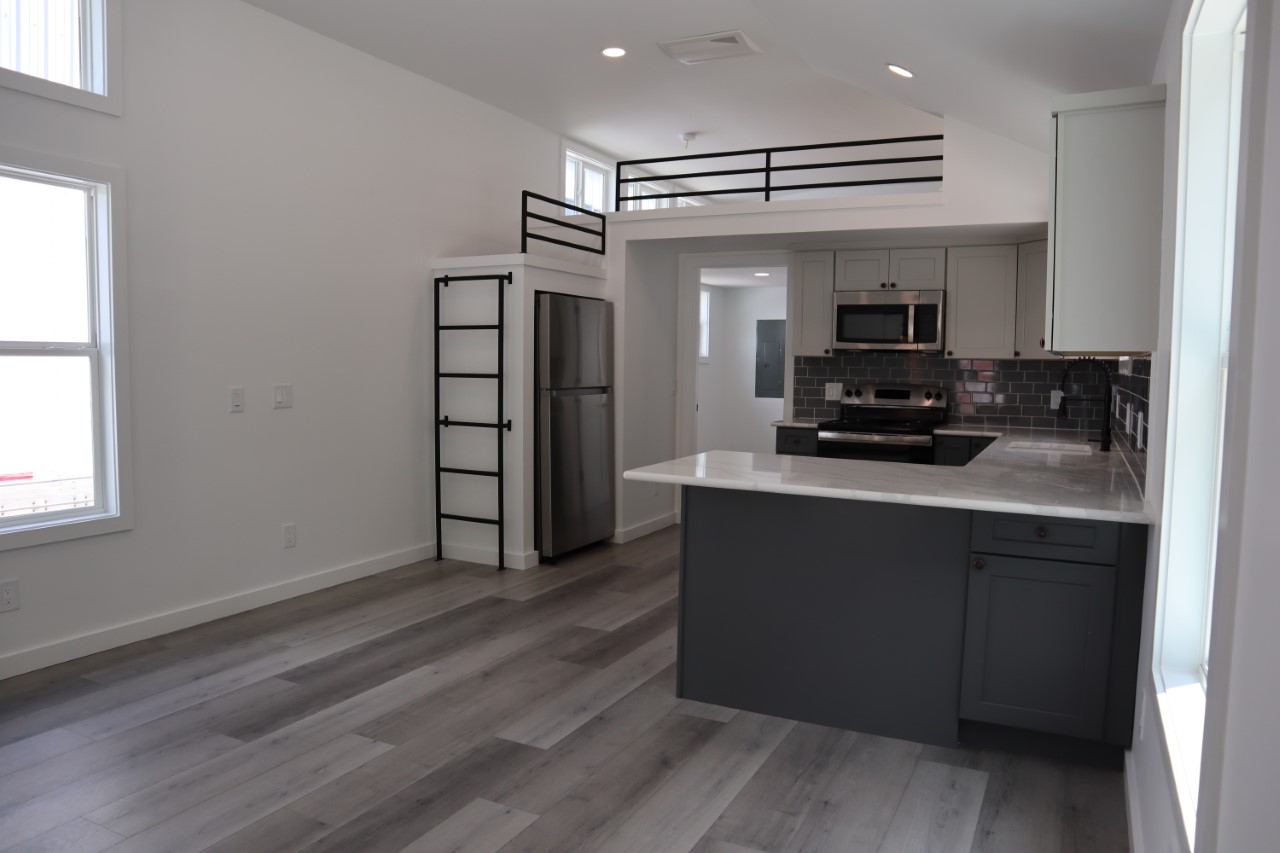 Modern kitchen with gray cabinets and stainless steel appliances. Loft above fridge adds space. Light gray wood floors and large windows enhance brightness.